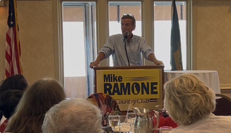 Mike Ramone speaking at a podium with a Mike Ramone sign, the American flag and the Delaware flag behind him.