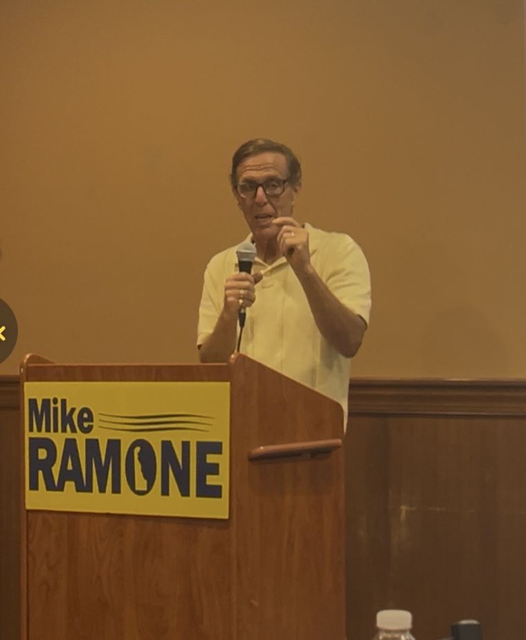 Mike Ramone speaking into a microphone behind a podium bearing his campaign sign.